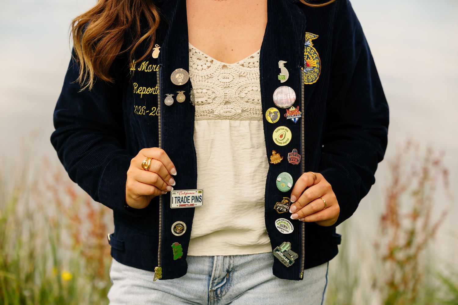 Woman displaying a navy blue jacket adorned with colorful enamel pins and badges, wearing a white lace top and jeans.