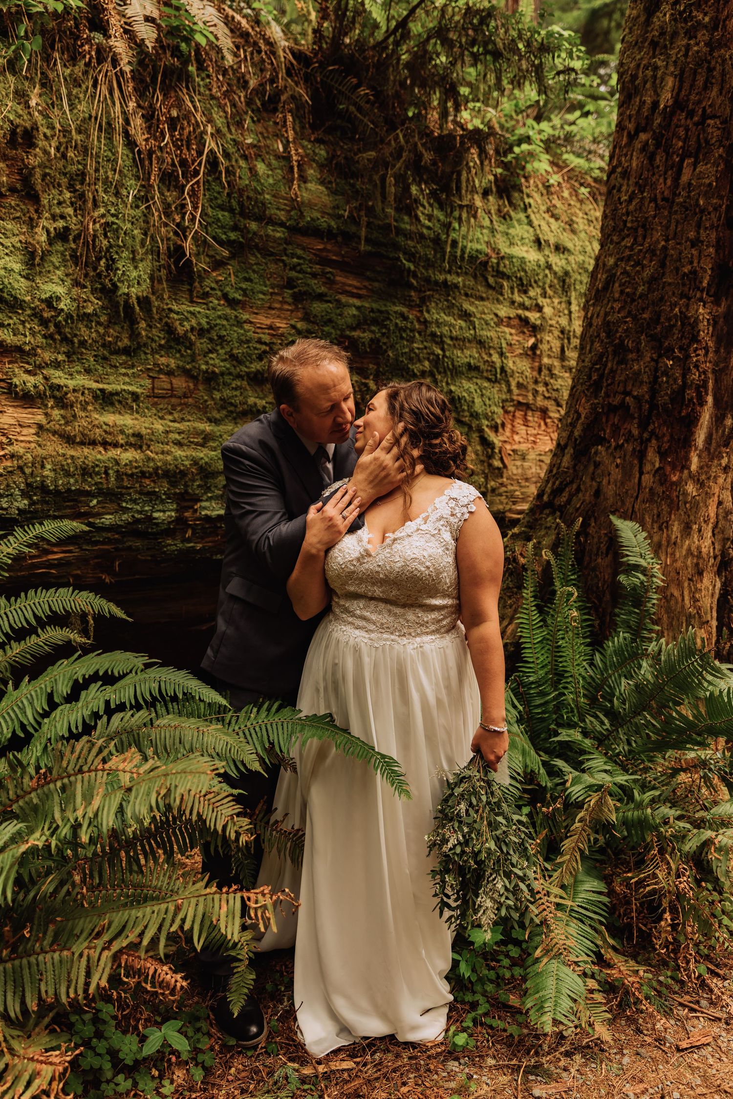 A wedding couple shares intimate moments among towering redwood trees and lush ferns in a forest setting.