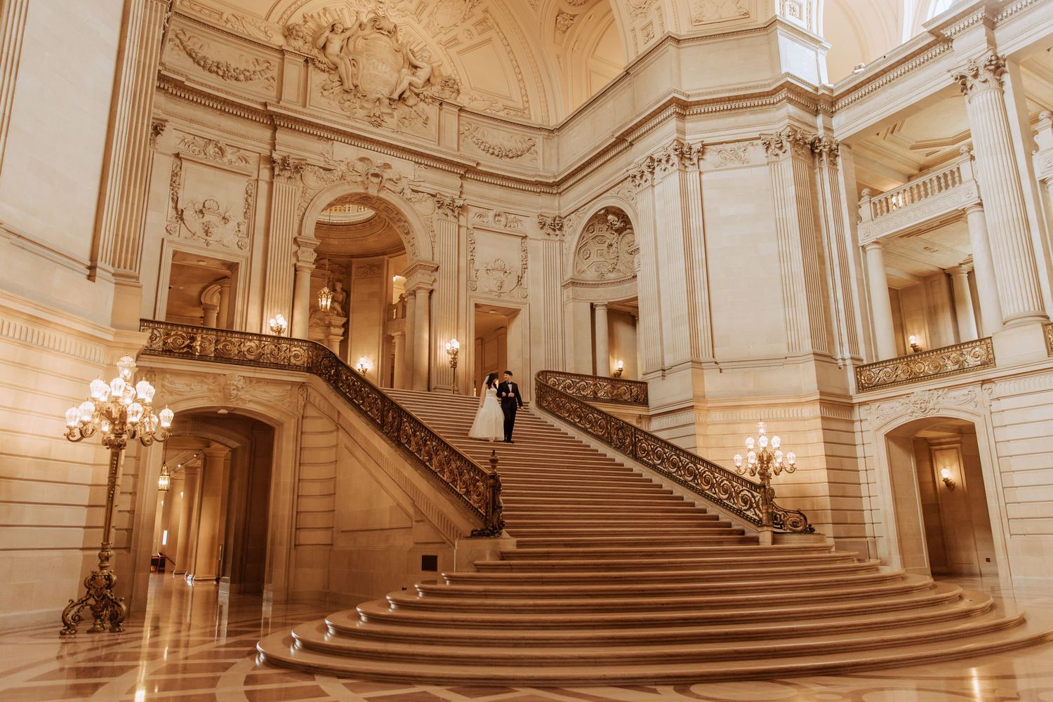 Ornate grand staircase in cream-colored baroque palace with crystal chandeliers.