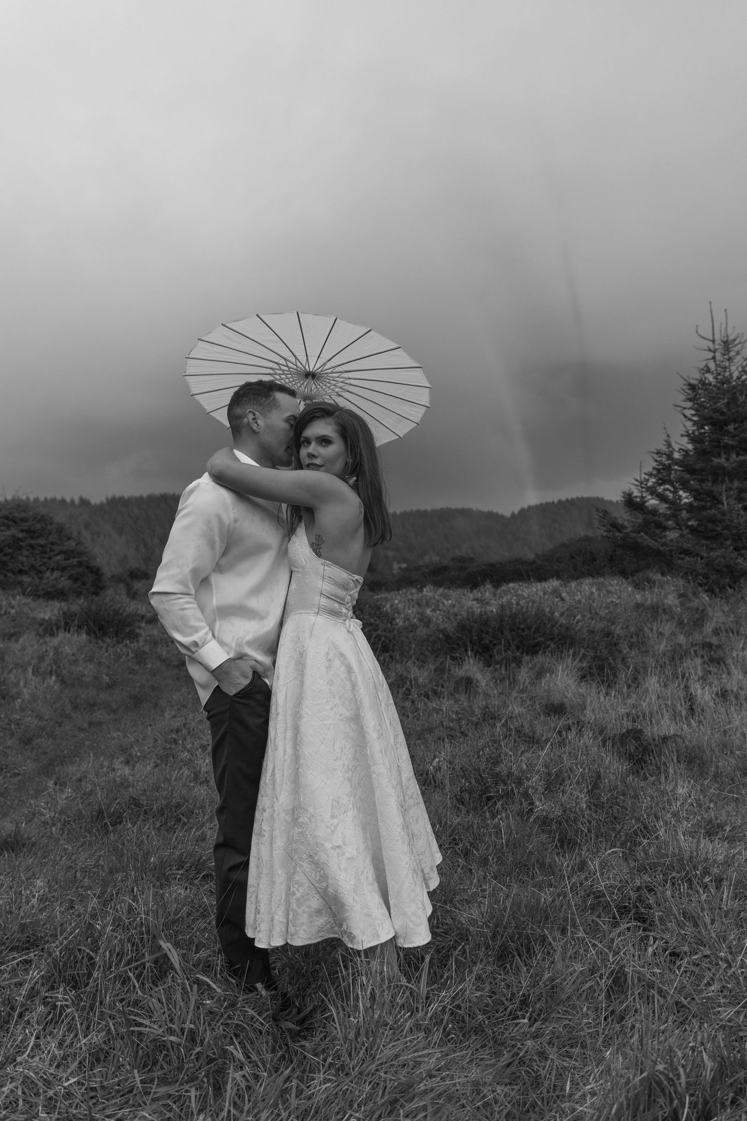 A couple poses with a white parasol against a moody grey coastal landscape.