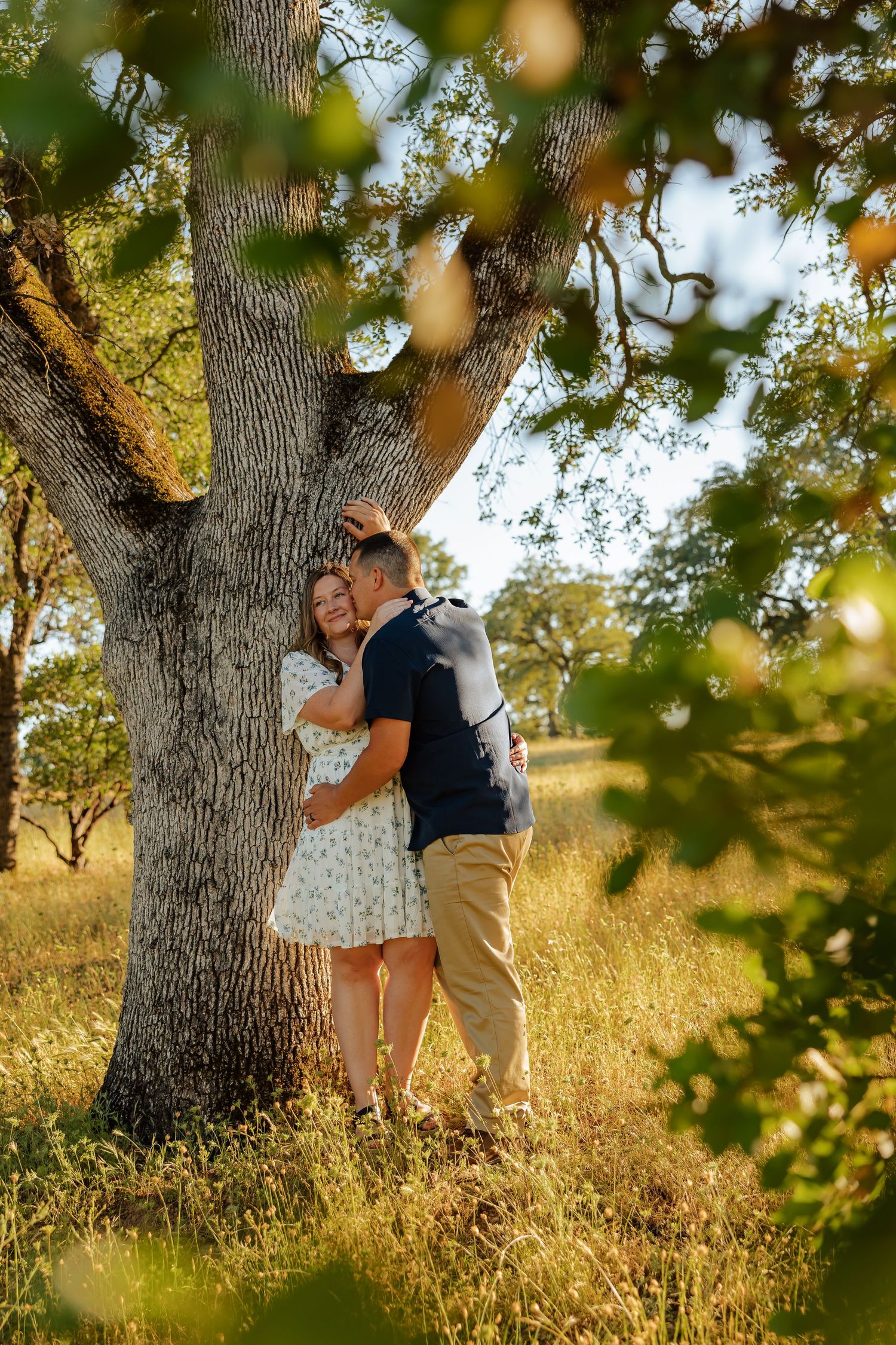 Couple embracing against a large oak tree in golden sunlight, woman in floral dress and man in navy shirt.