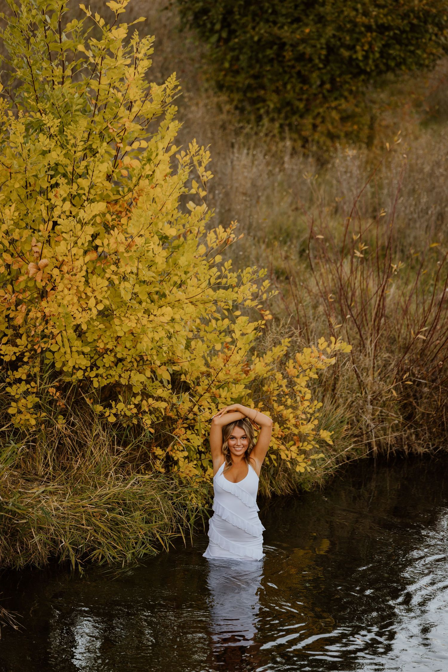 Woman in white tank top stands in water beside golden autumn tree at sunset.