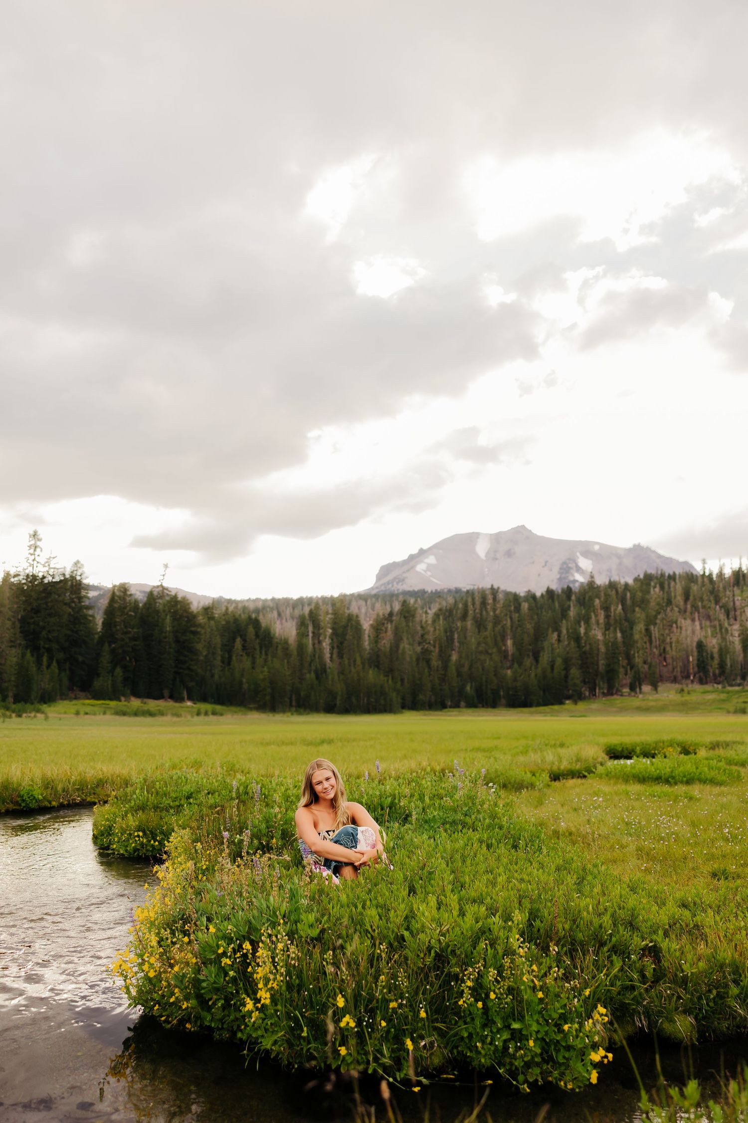 Woman sitting by a stream in a lush green mountain meadow with wildflowers and forested peaks under cloudy skies.