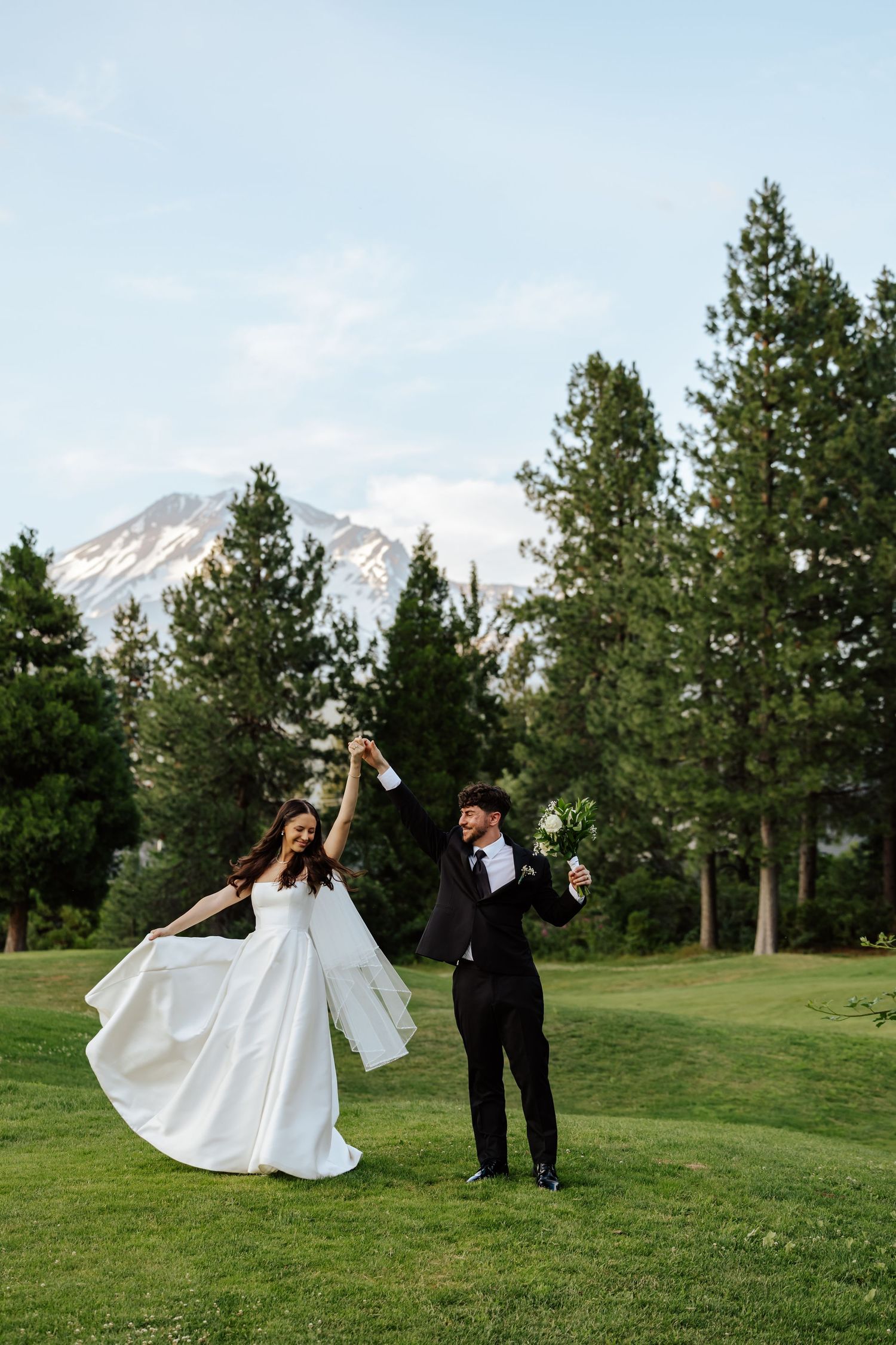 Joyful bride and groom celebrating on a green lawn with snow-capped mountains and pine trees behind them.