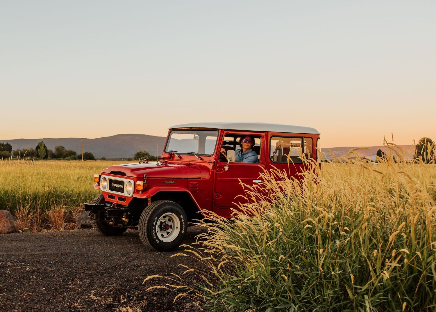 Red Toyota Land Cruiser parked in field during golden hour with mountains in background.