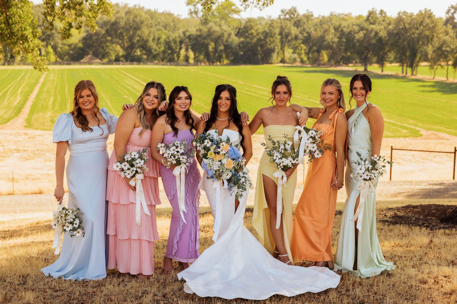 A wedding party in pastel dresses poses together in a sunlit outdoor setting.