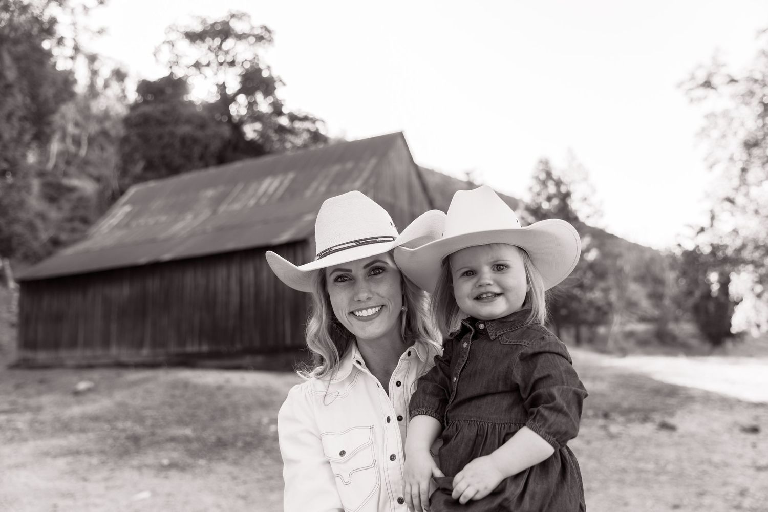 Family photoshoot outdoors with rustic barn and cowboy hats in black and white style.