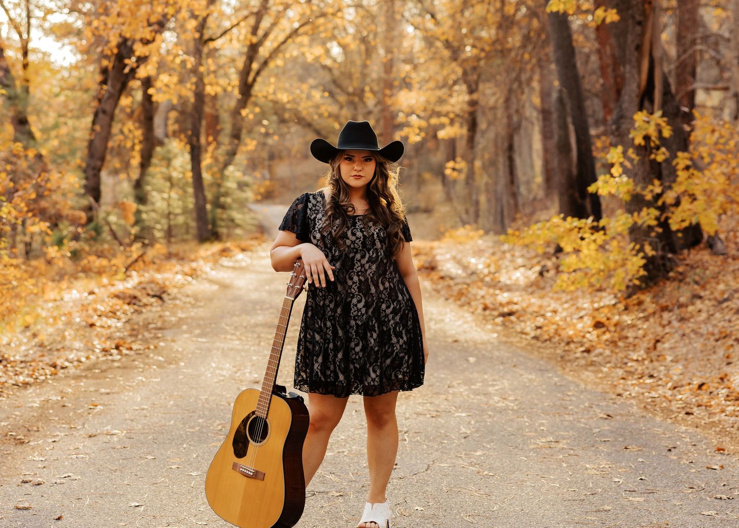 Woman in black dress poses with acoustic guitar on autumn forest path.