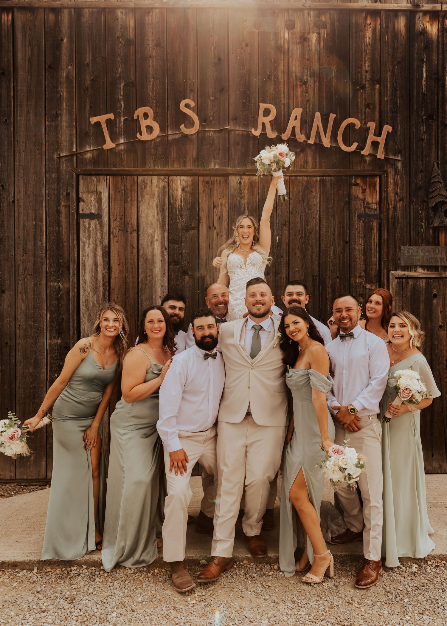 Wedding party poses casually against rustic wooden barn wall wearing sage green dresses and light colored suits.
