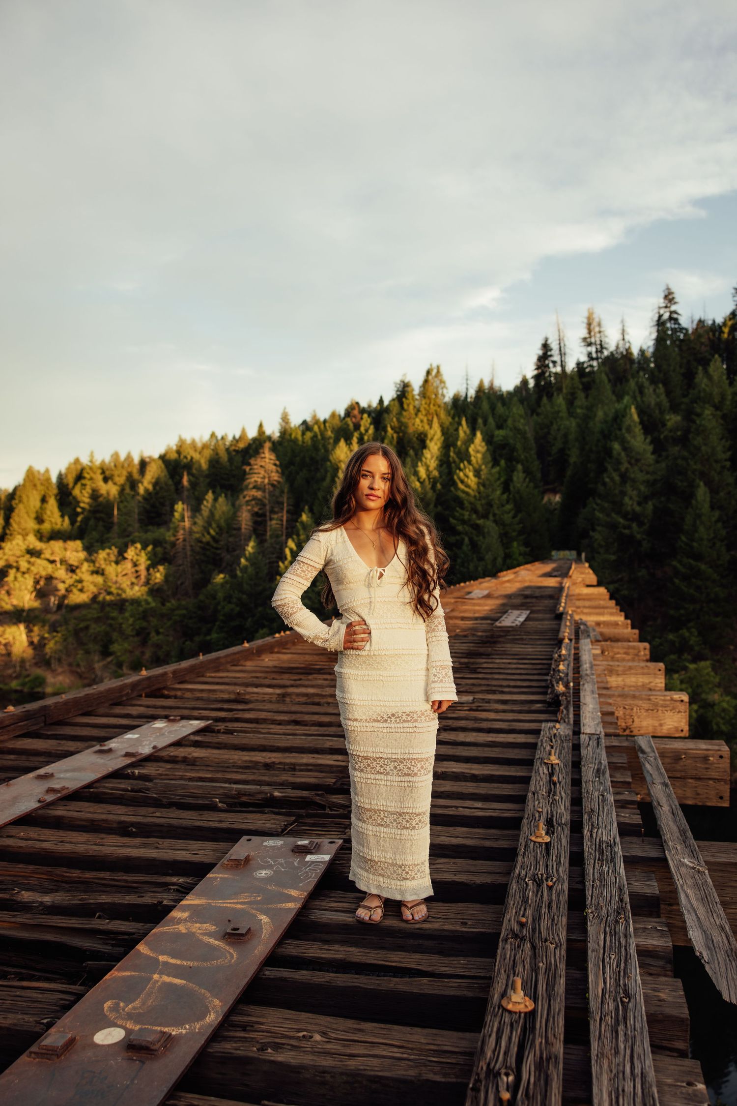 White lace dress photoshoot on rustic wooden bridge with forest backdrop.