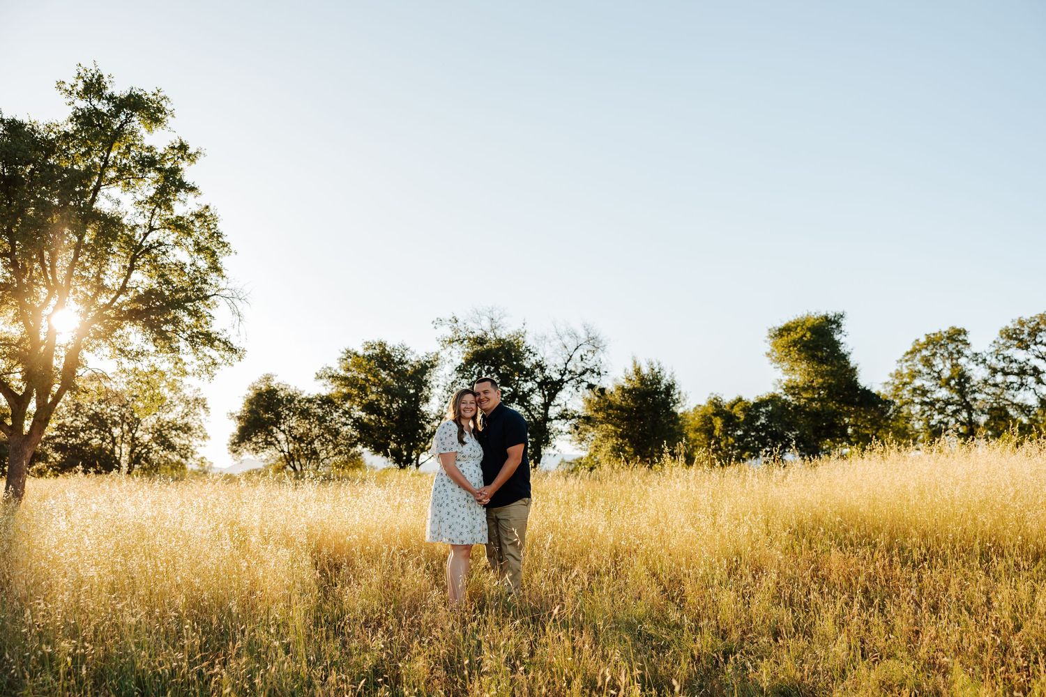 Couple sharing a romantic moment in a vast golden field with oak trees and bright sunlight behind them.