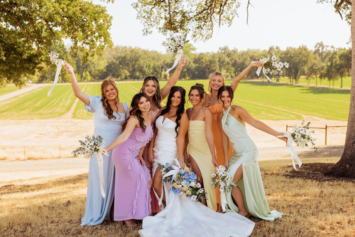 Bridesmaids in pastel dresses pose together on a grassy hillside during golden hour.