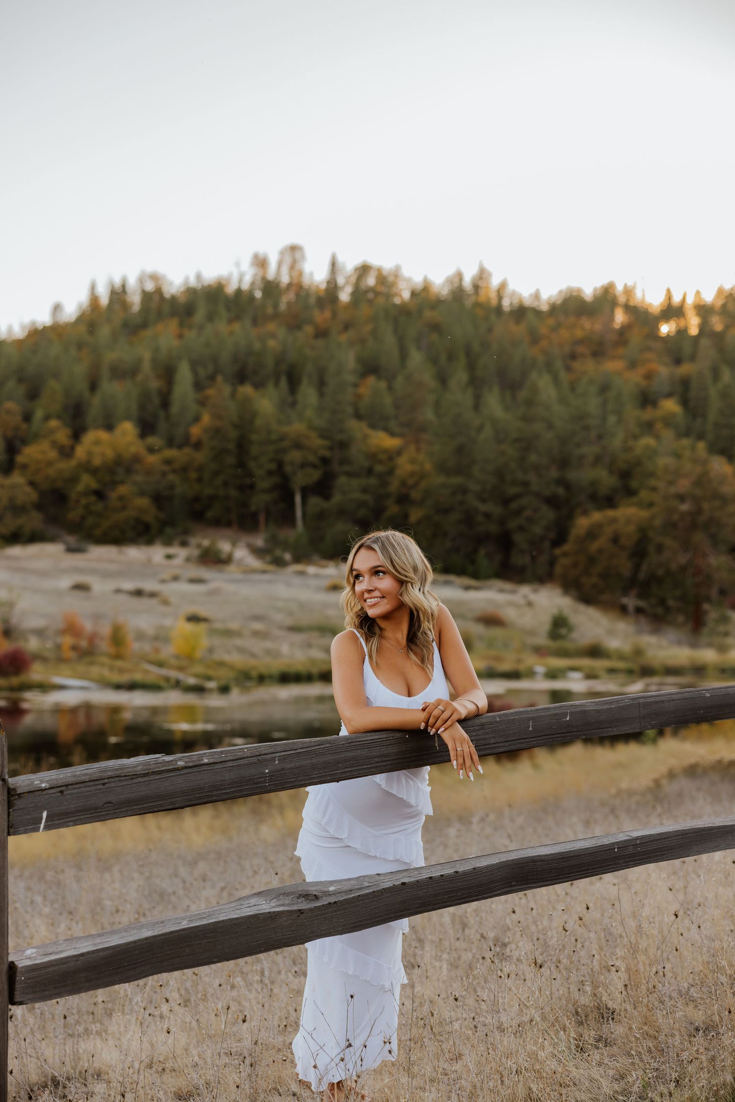 Woman in white dress leans on wooden fence during sunset with autumn landscape behind.