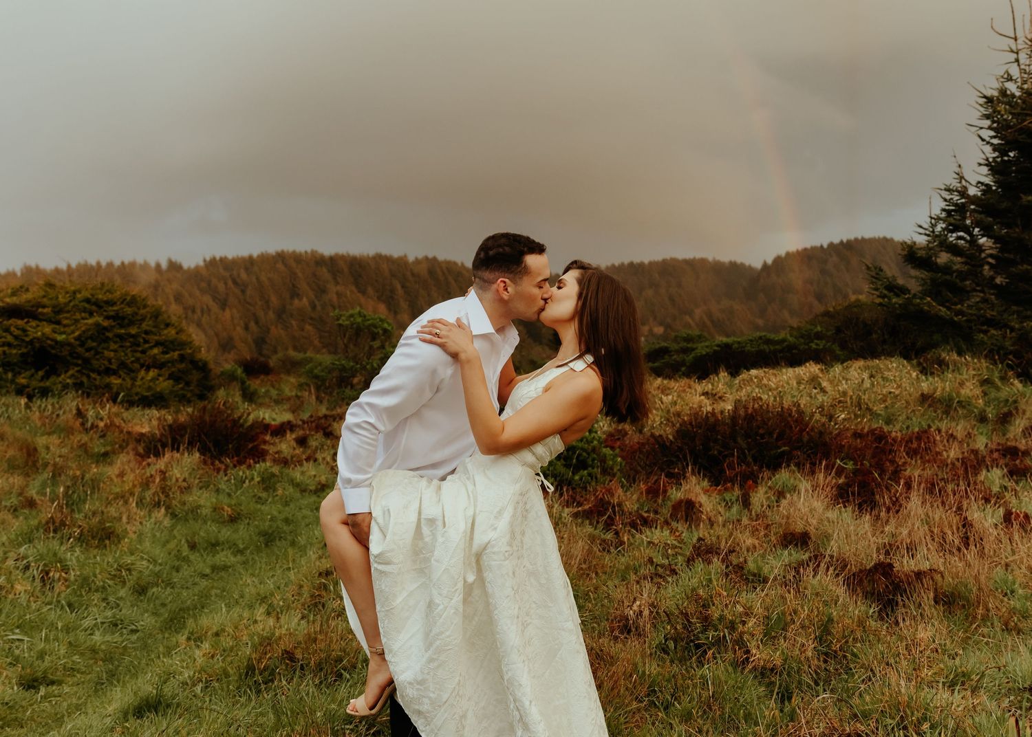 A couple in formal wear embraces on a scenic coastal hillside during golden hour.
