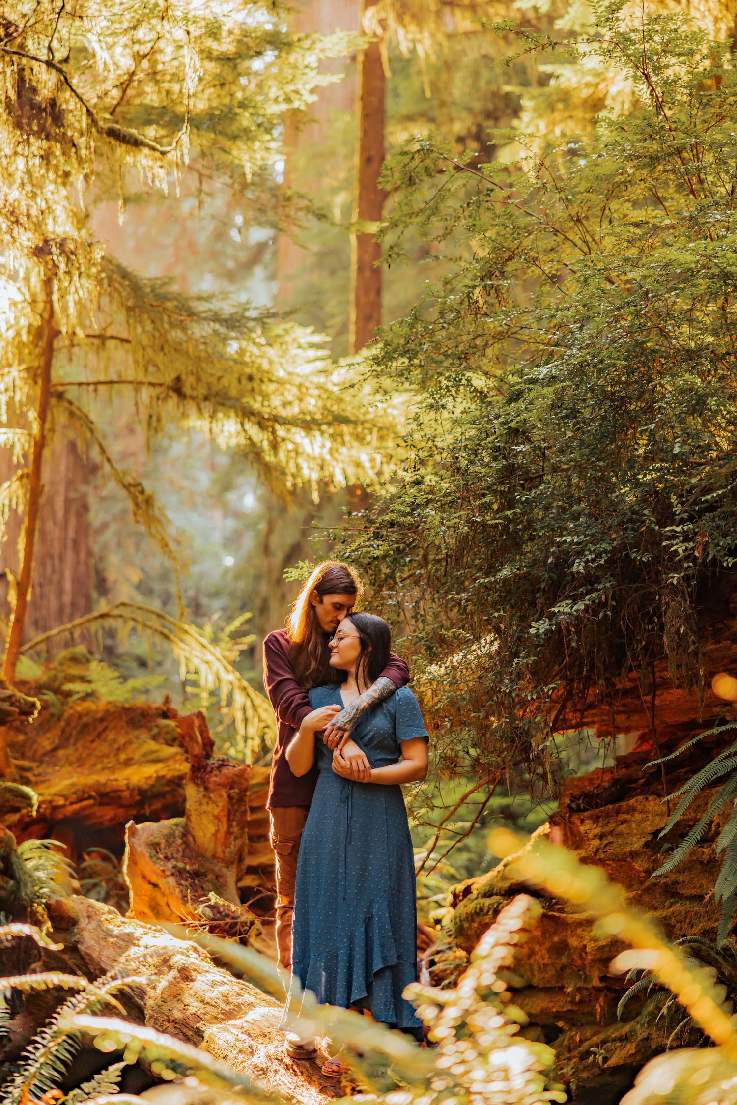 A tender embrace photographed in a sunlit forest with golden light filtering through the trees.