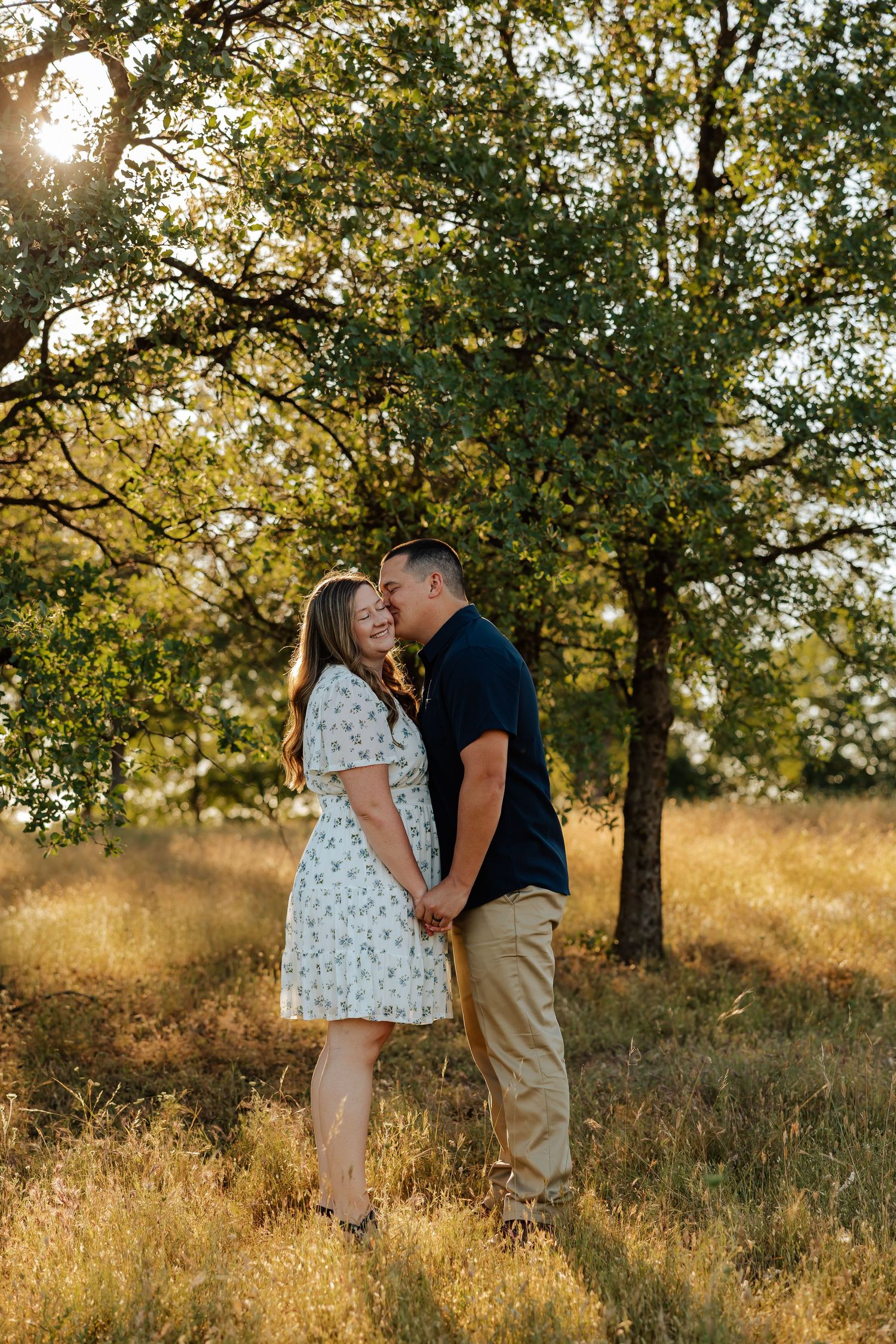 Couple holding hands and almost kissing under a sunlit tree in a golden meadow, woman in floral dress.