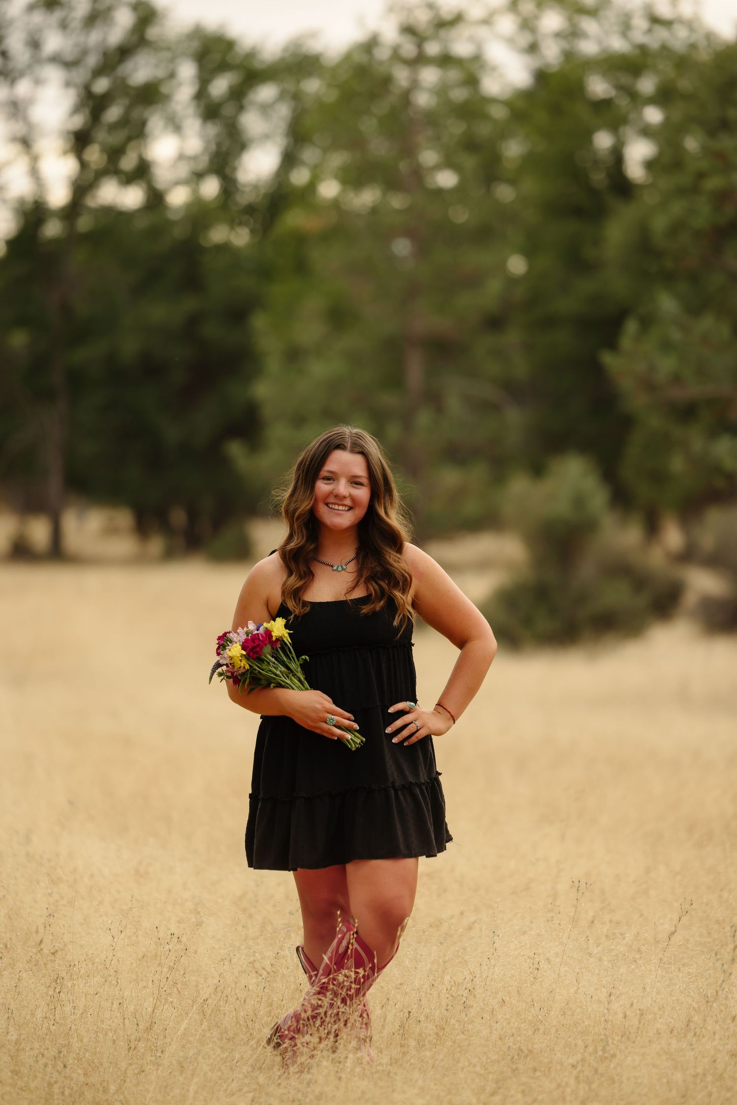 Young woman in black dress and cowboy boots holding wildflowers while standing in a golden field with trees behind her.