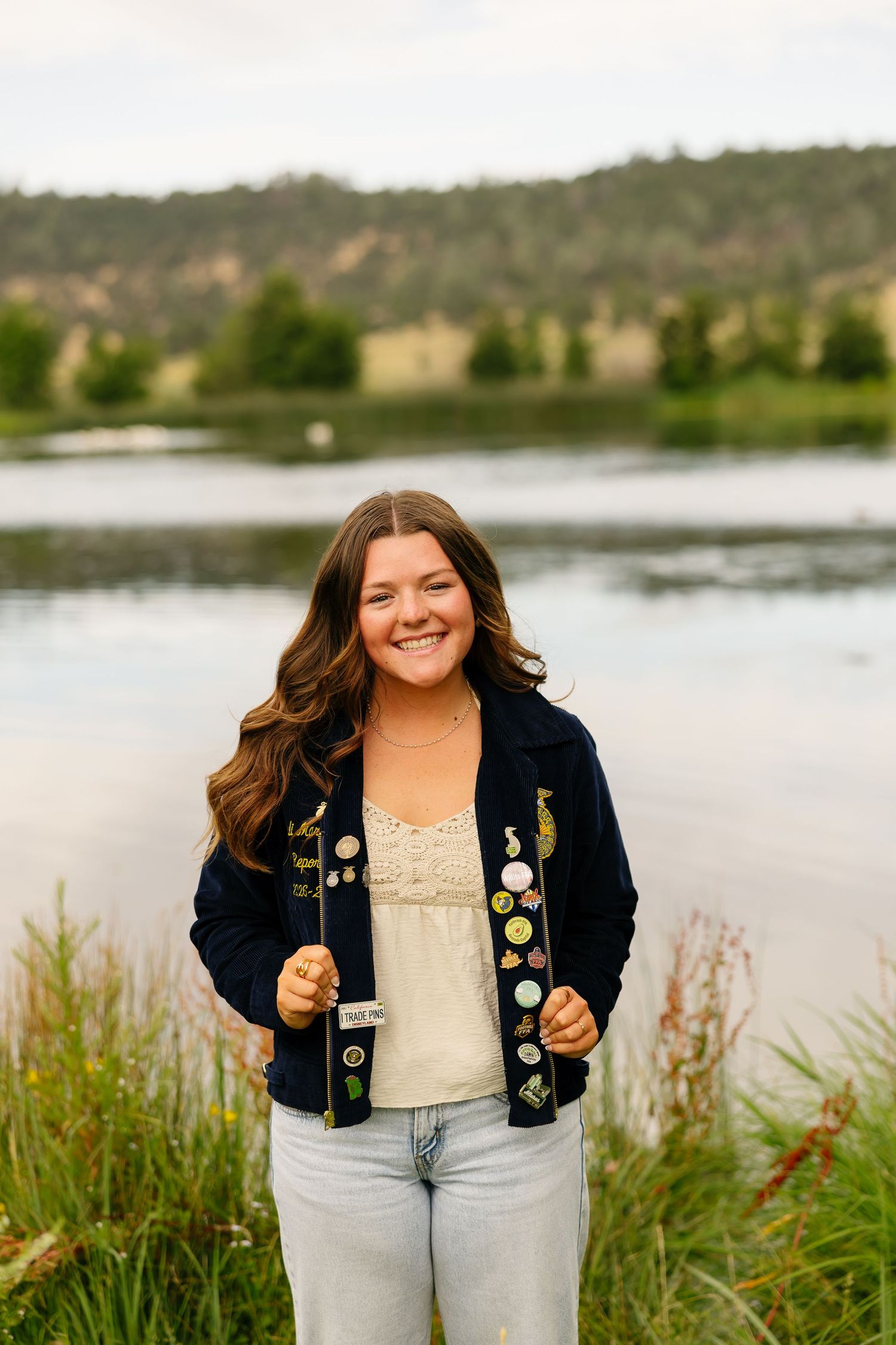 Smiling young woman in a navy cardigan with pins stands by a serene lake with green hills in the background.