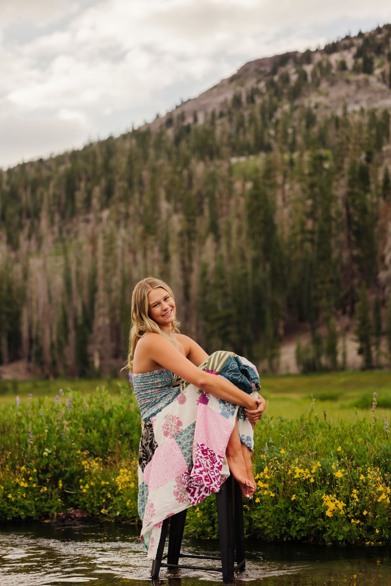 Smiling blonde woman wrapped in colorful quilt standing in mountain stream with wildflowers and pine trees.