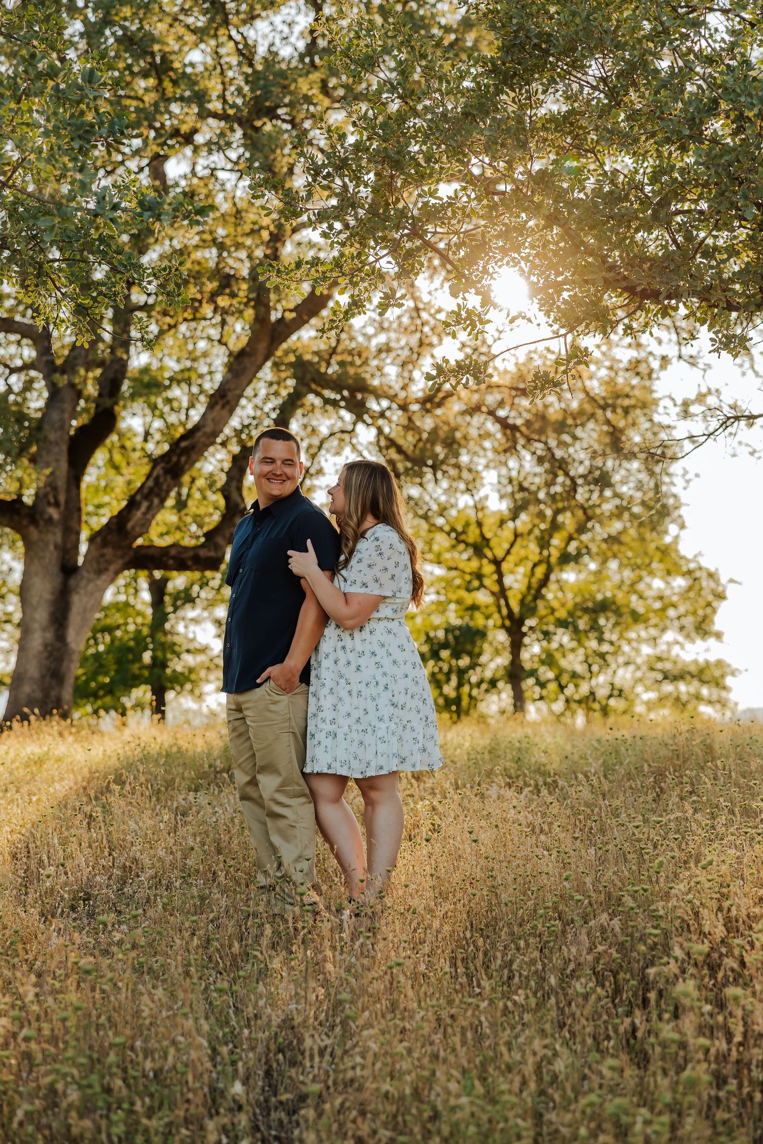 Couple embracing in golden sunlit field beneath oak trees, woman in floral dress leaning toward man in navy shirt.