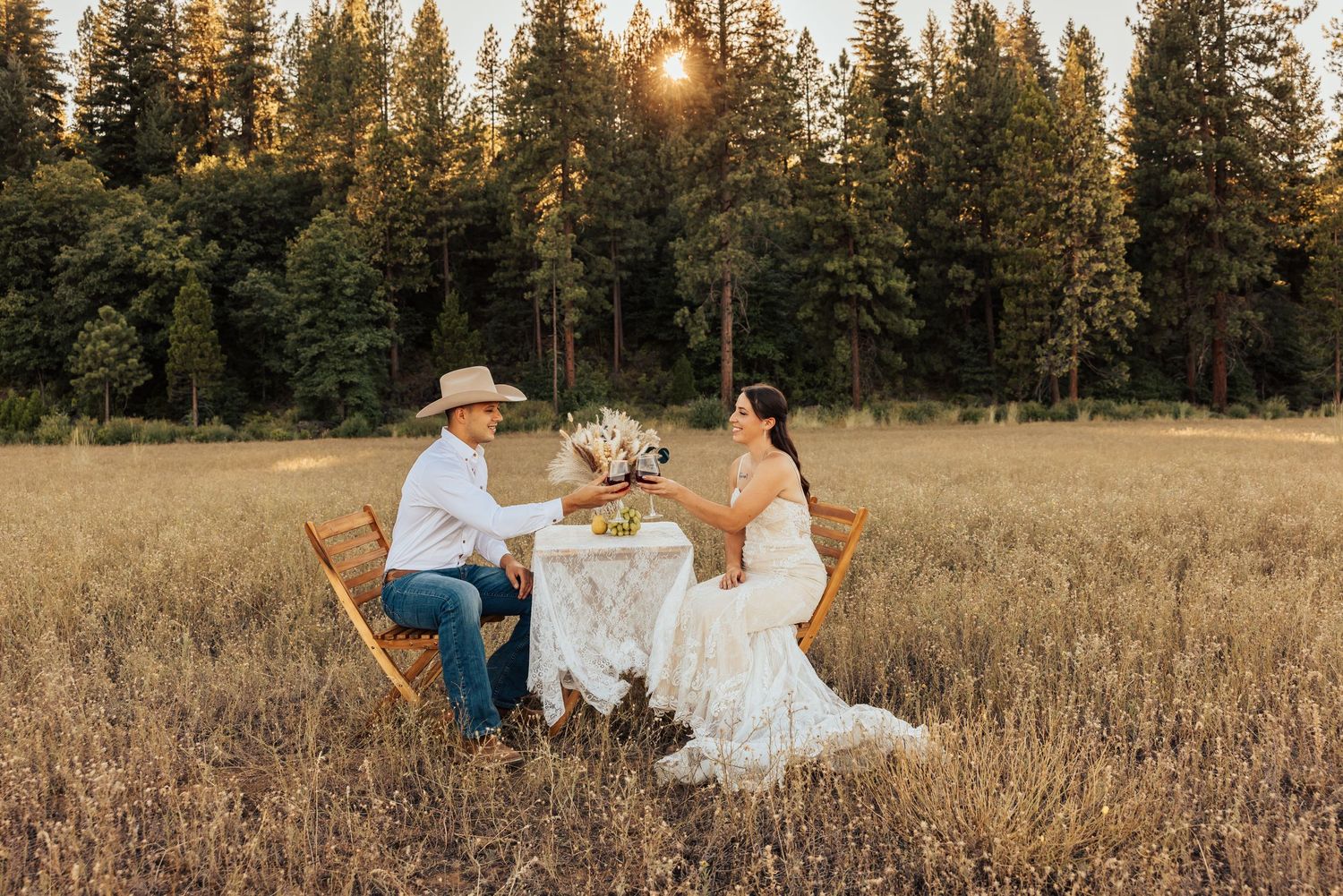 A rustic outdoor wedding reception scene with a couple dining at sunset in a golden meadow.