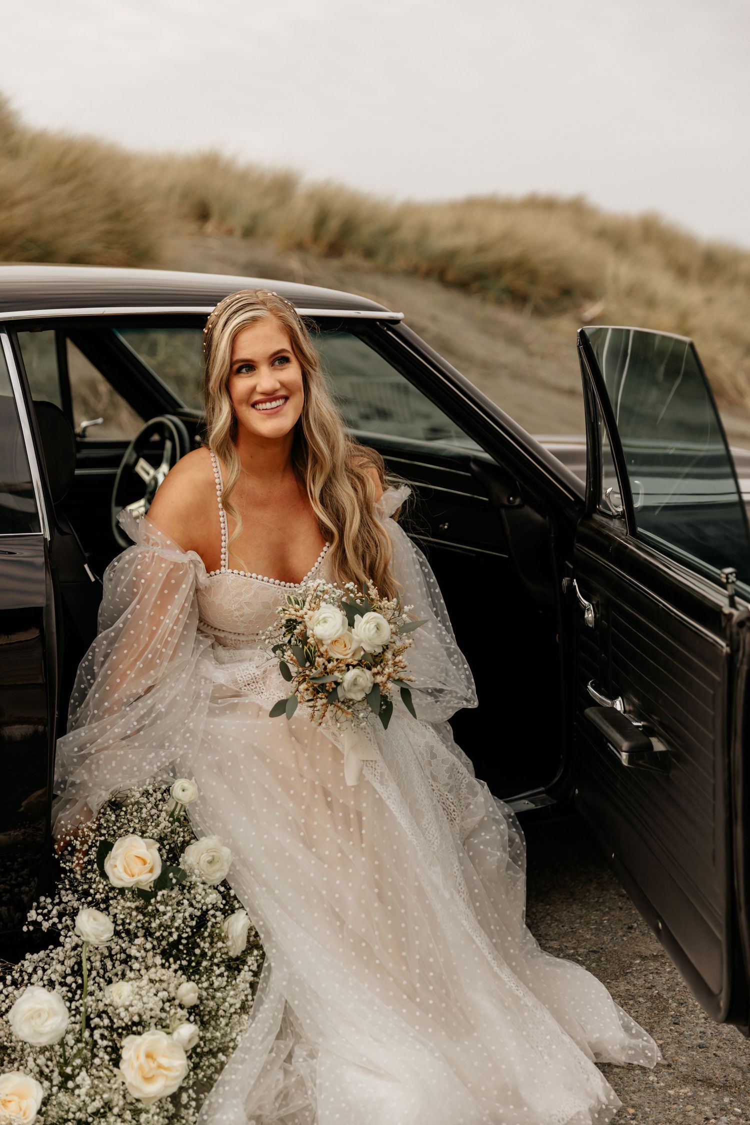 Joyful bride in white lace gown sitting in vintage car with floral bouquet.