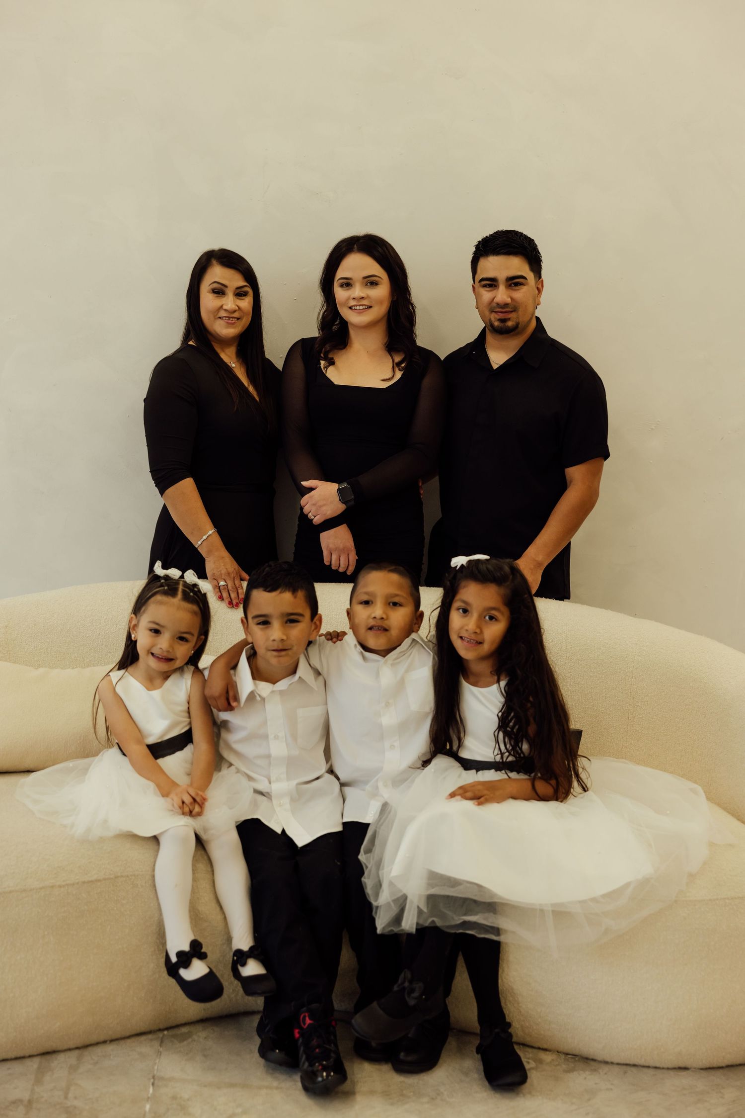 Children in white formal attire pose with adults in black on a light-colored couch.
