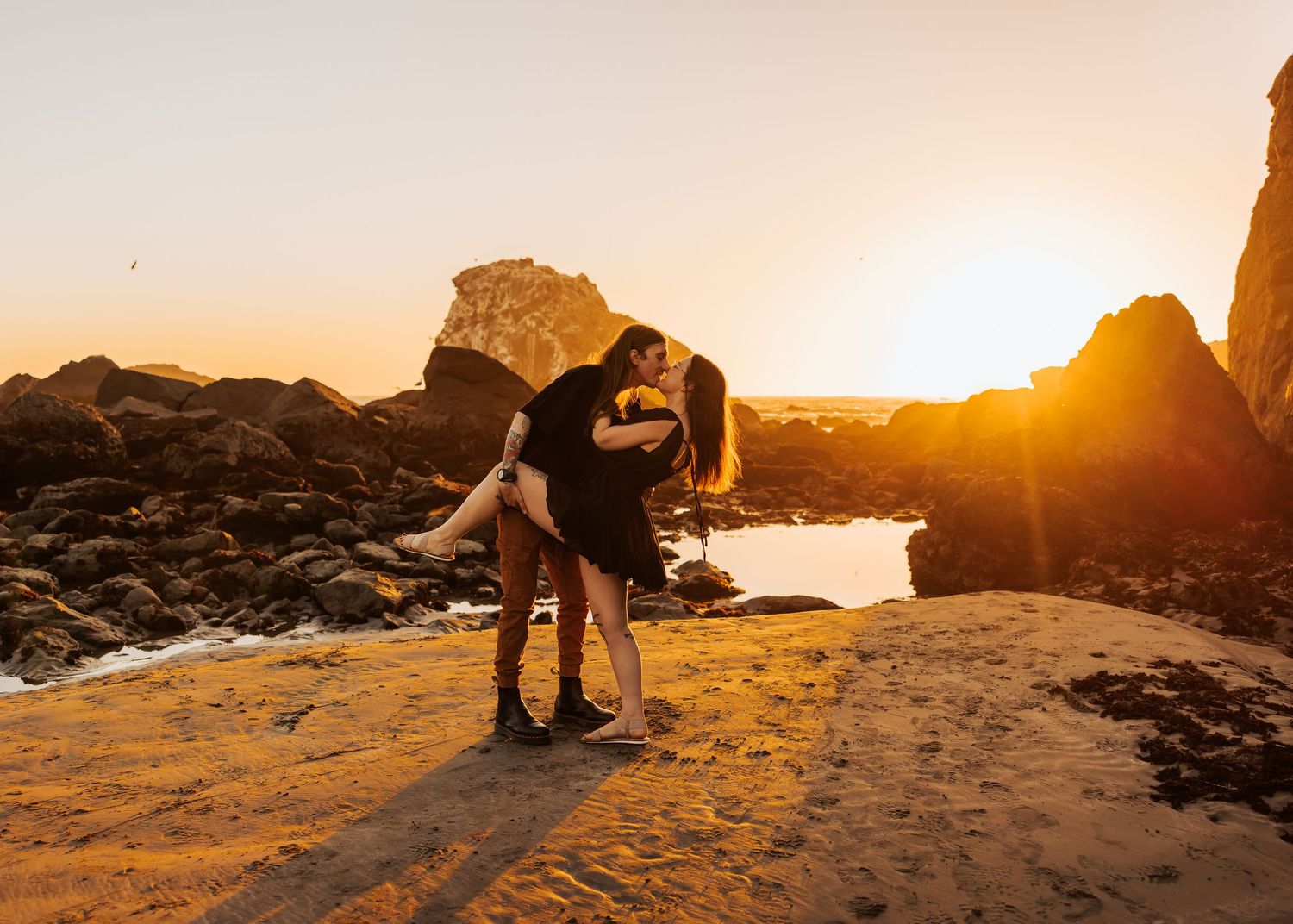 Silhouetted couple sharing romantic moment on rocky beach during spectacular sunset.