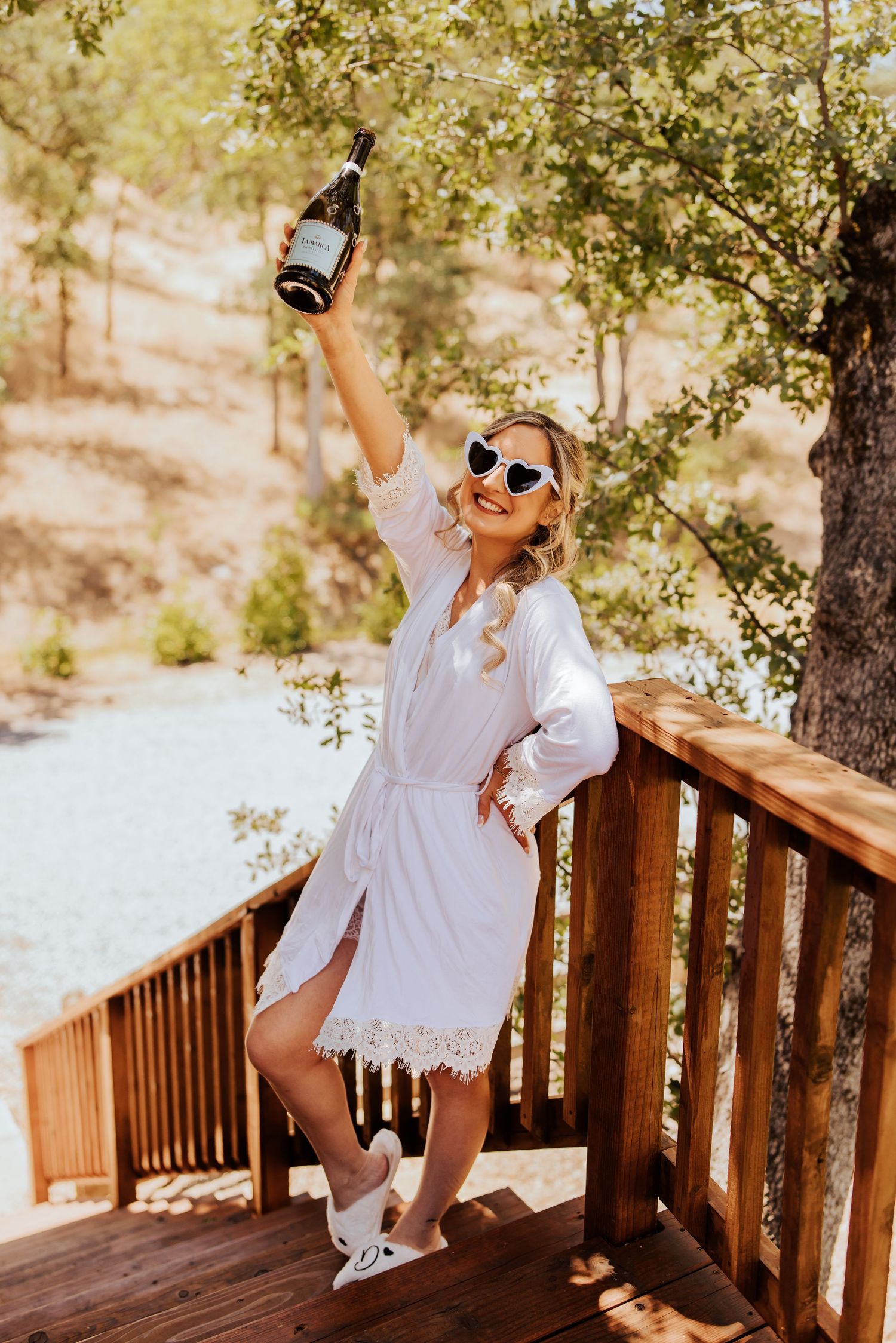 Bridal preparation photos on wooden deck wearing white lace robe and sunglasses.