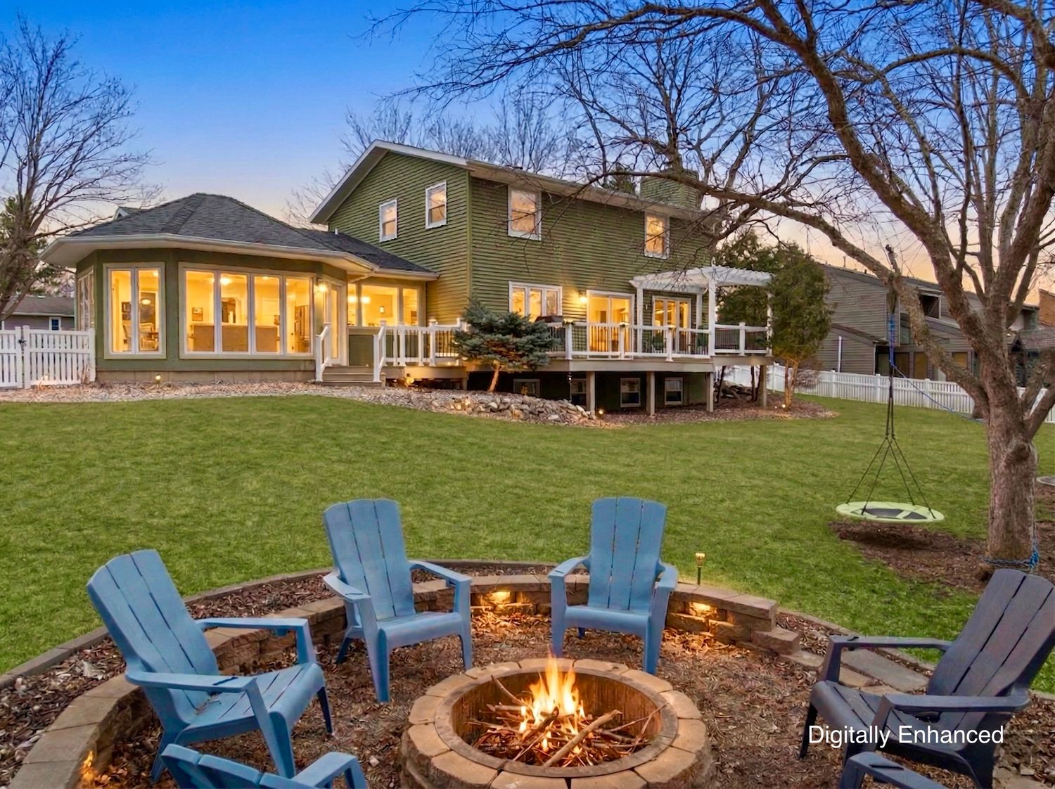 Backyard patio with blue Adirondack chairs around a stone fire pit, lush lawn, and large green home with deck at dusk.