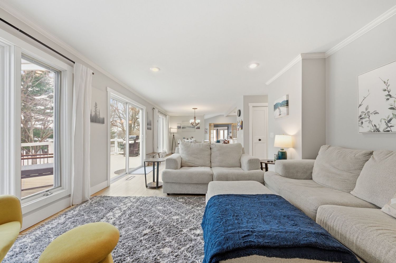 Bright living room with white sofas, blue throw, large windows, hardwood floors, and open layout leading to dining area.