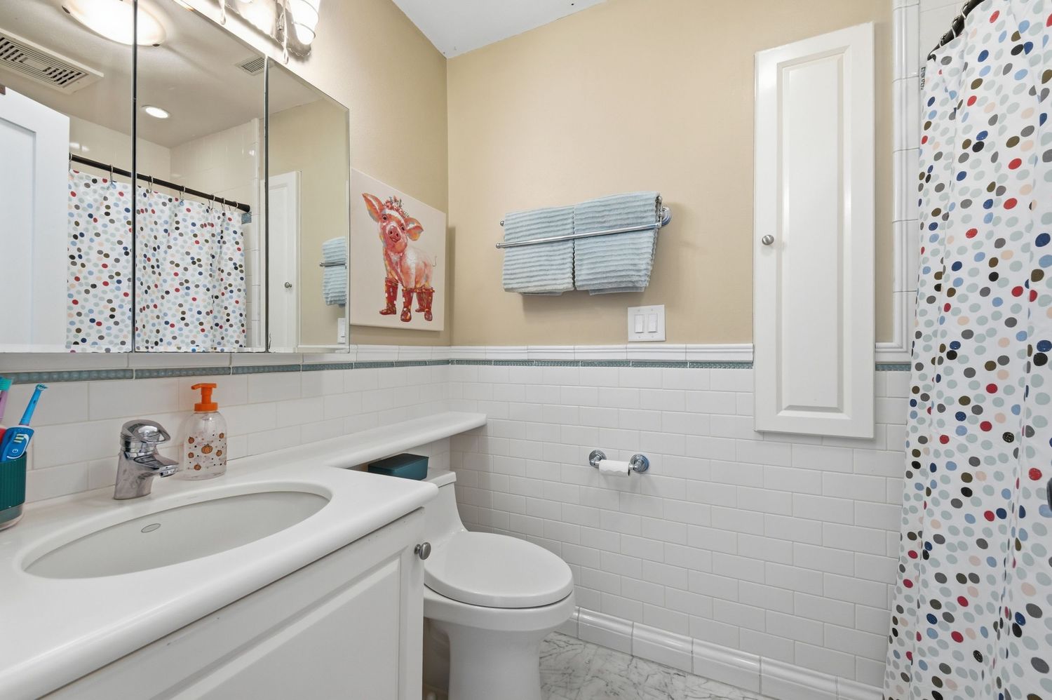 Bright white bathroom with tile wainscoting, vanity with mirror, toilet, medicine cabinet, and colorful polka dot shower curtain.