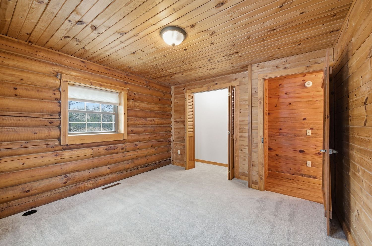 Empty bedroom with wood paneled walls, carpet flooring, and natural light from a window in a rustic cabin interior.
