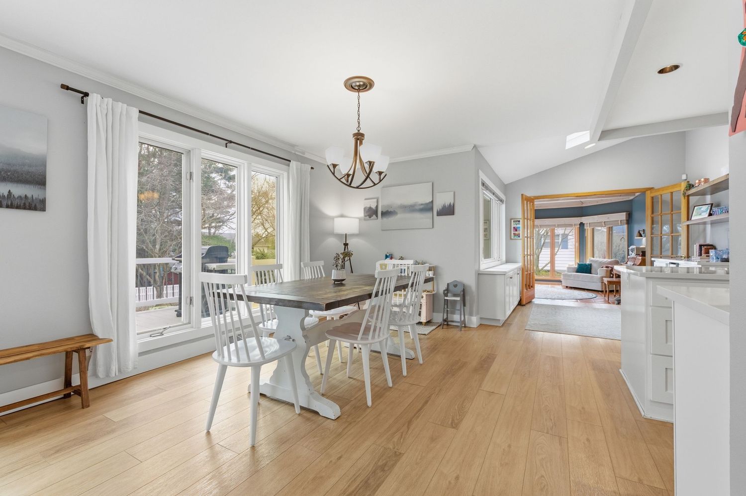 Bright dining room with hardwood floors, white farmhouse table, chandelier, and large windows overlooking a wooded backyard.