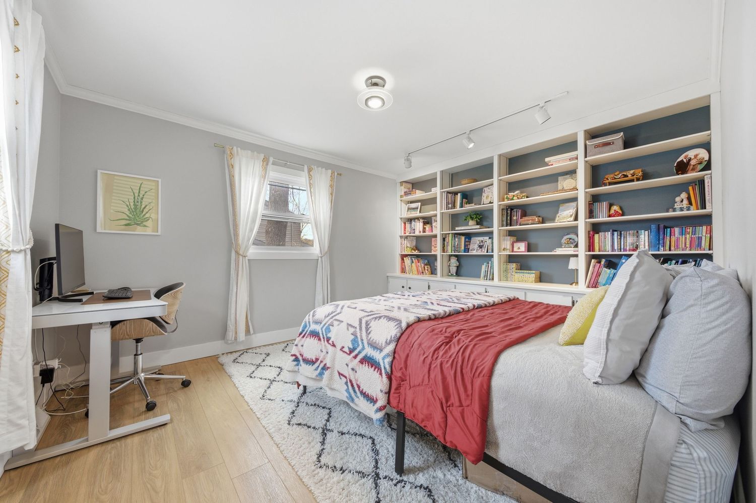 Bright bedroom with a queen bed, red throw blanket, built-in bookshelves, hardwood floors, and a white desk by the window.