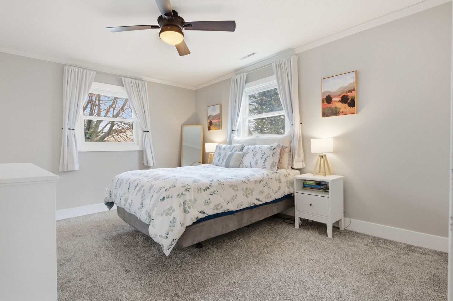 Bright bedroom with ceiling fan, plush carpet, queen bed with floral bedding, white nightstand, and two windows with curtains.
