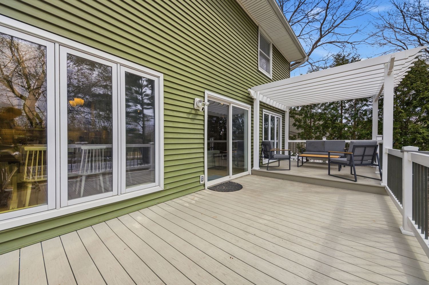 Spacious outdoor deck with gray composite flooring, white pergola seating area, and large sliding glass doors on a green-sided home.