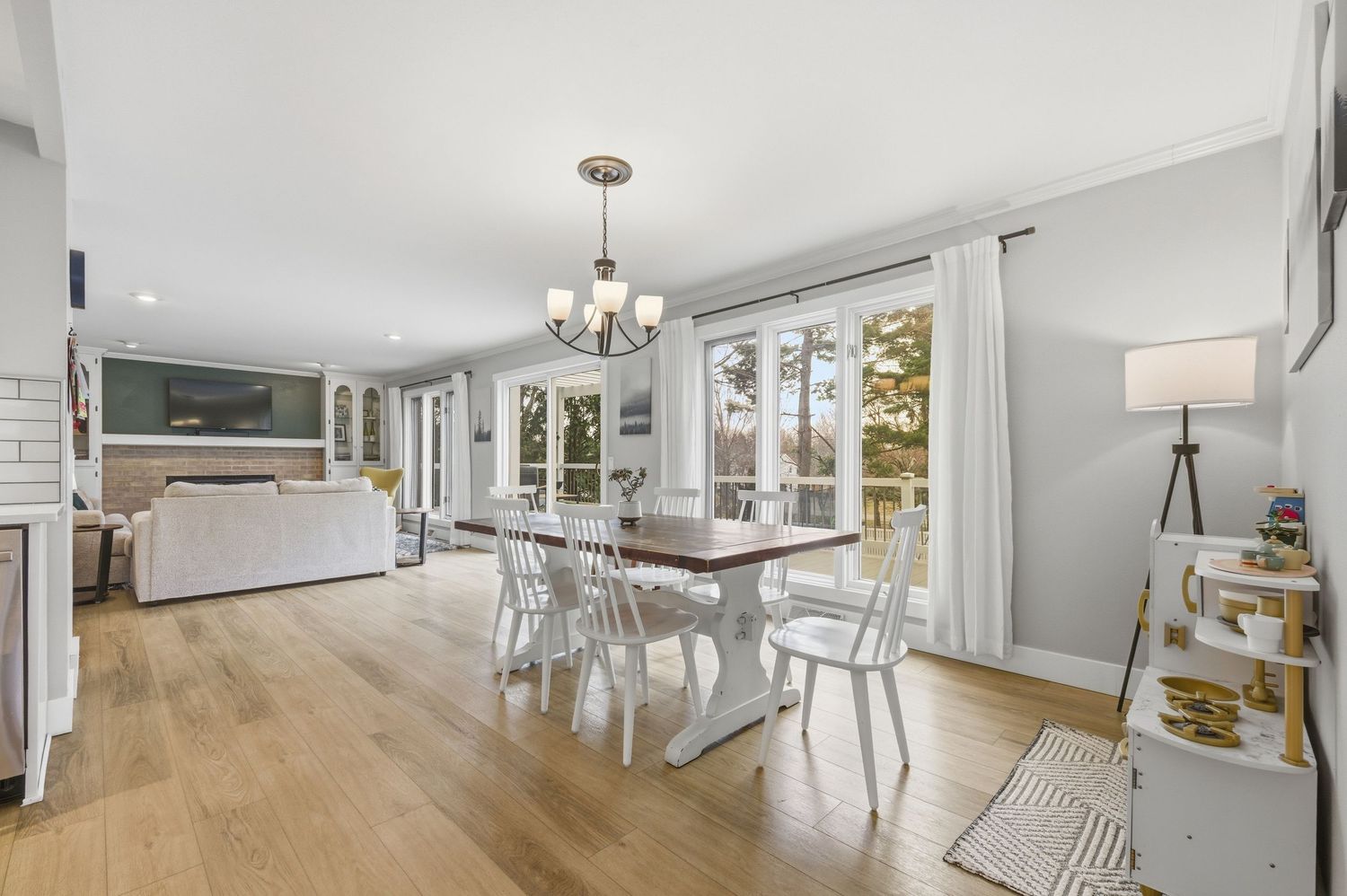 Bright open-plan dining room with white chairs, wood table, hardwood floors, chandelier, and large windows overlooking outdoor views.