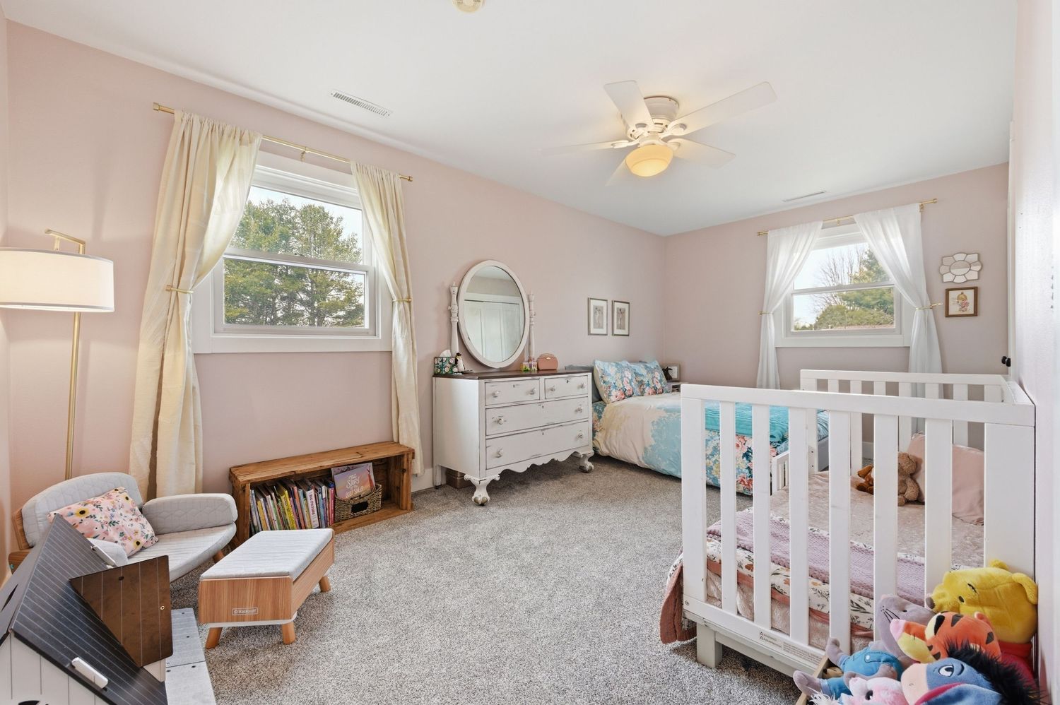 Pink nursery with white crib, dresser, ceiling fan, and two windows with curtains on carpeted floor.