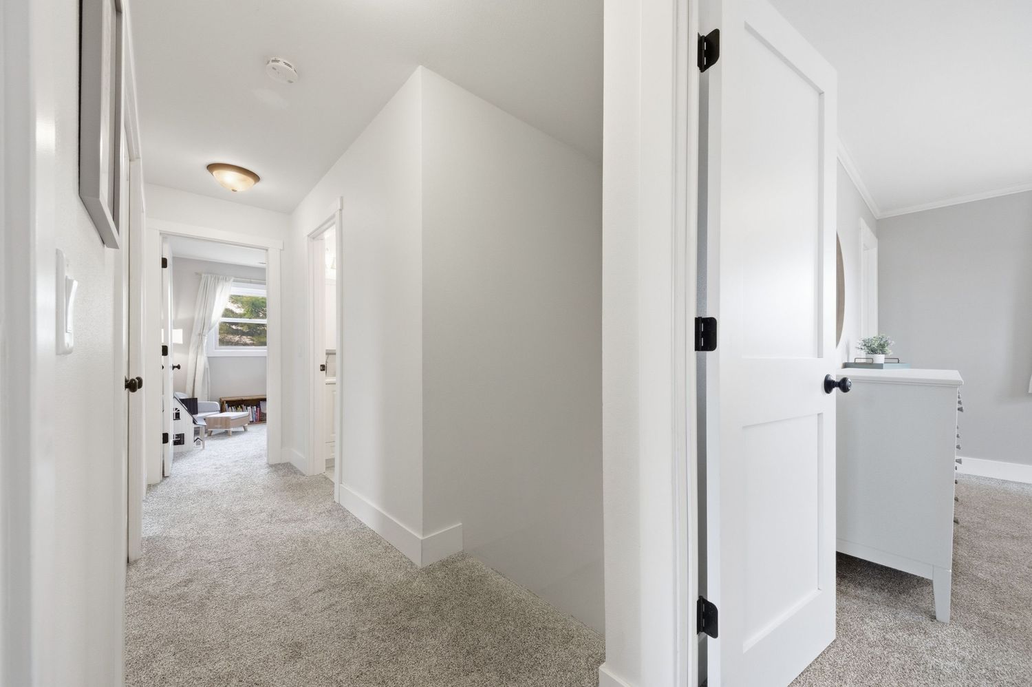 Bright hallway with light gray carpet, white walls, and white doors leading to a sunlit room beyond.