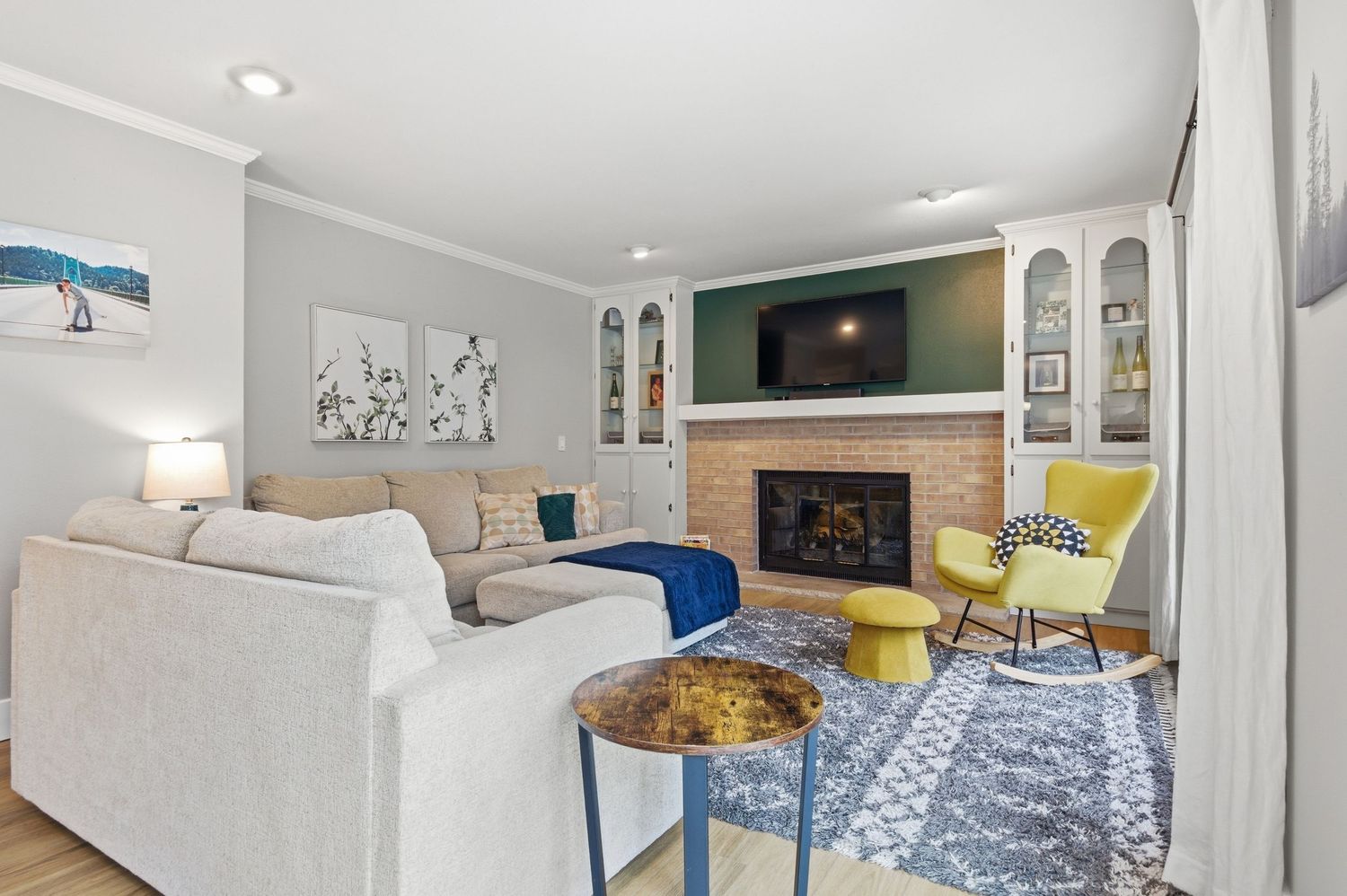 Cozy living room with white sofas, brick fireplace, built-in shelves, yellow accent chairs, and blue patterned rug.