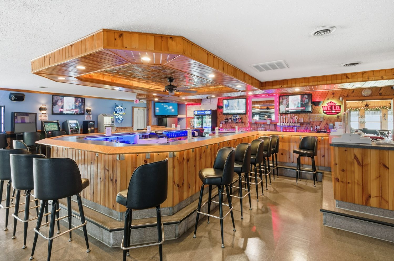 Long wooden bar counter with black barstools in a rustic restaurant interior with wood paneled walls and ceiling.