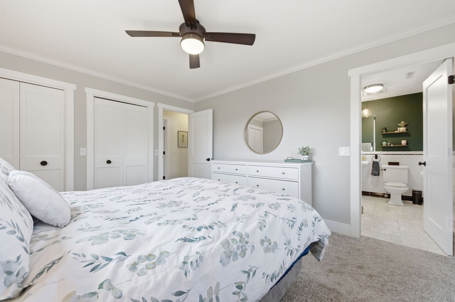 Bright bedroom with floral bedding, ceiling fan, white dresser, round mirror, and en-suite bathroom visible in background.