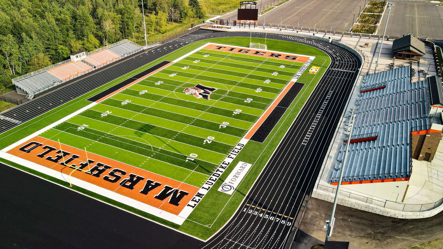 Marshfield Senior High School football stadium with bright green turf and orange endzones on a sunny day.