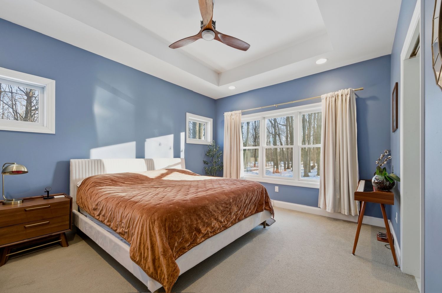Modern bedroom with blue walls, white trim, ceiling fan, and large windows showcasing natural light and brown bedding.