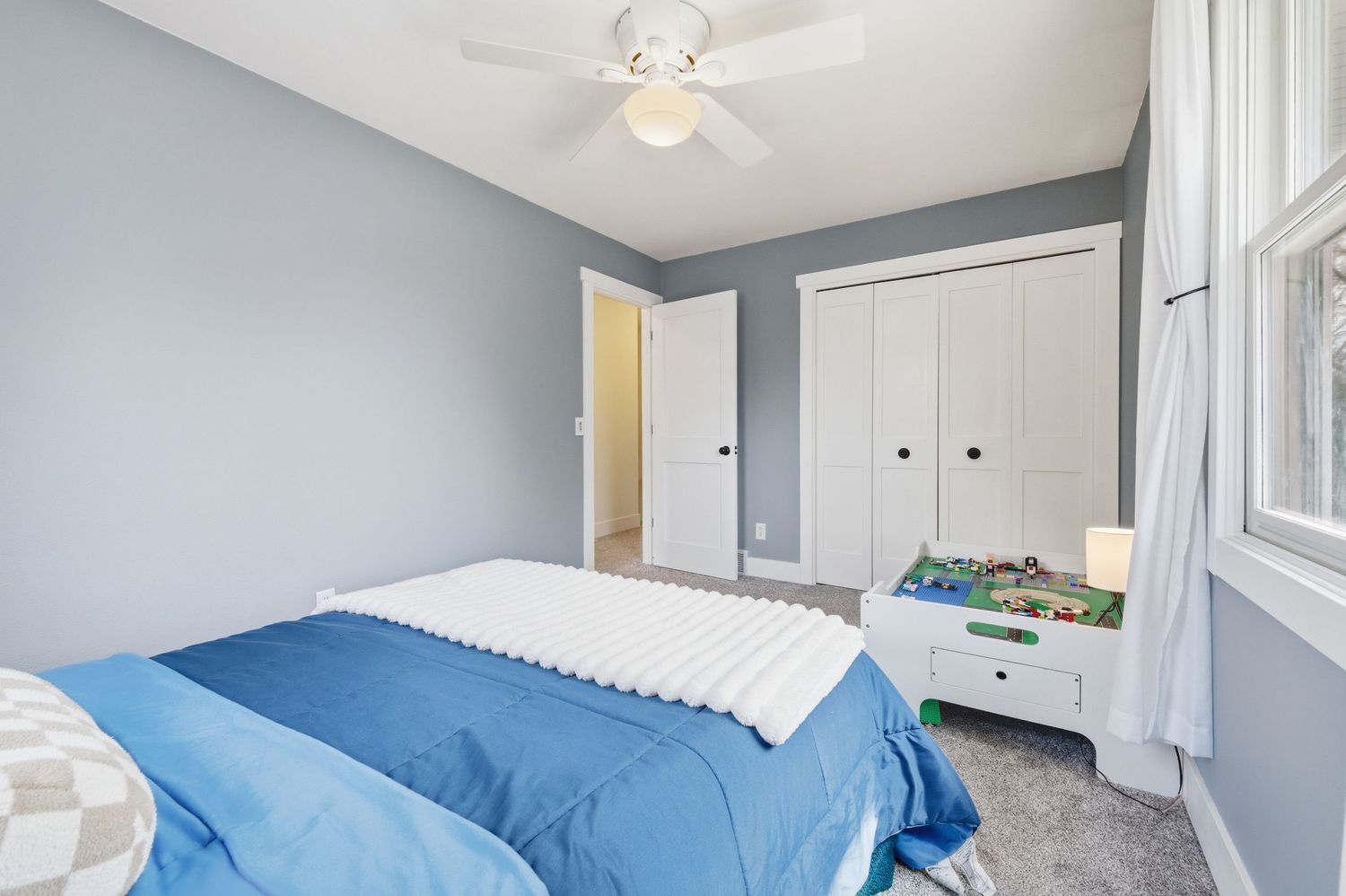 Cozy bedroom with blue bedding, white closet doors, ceiling fan, and gray walls in a well-lit space.