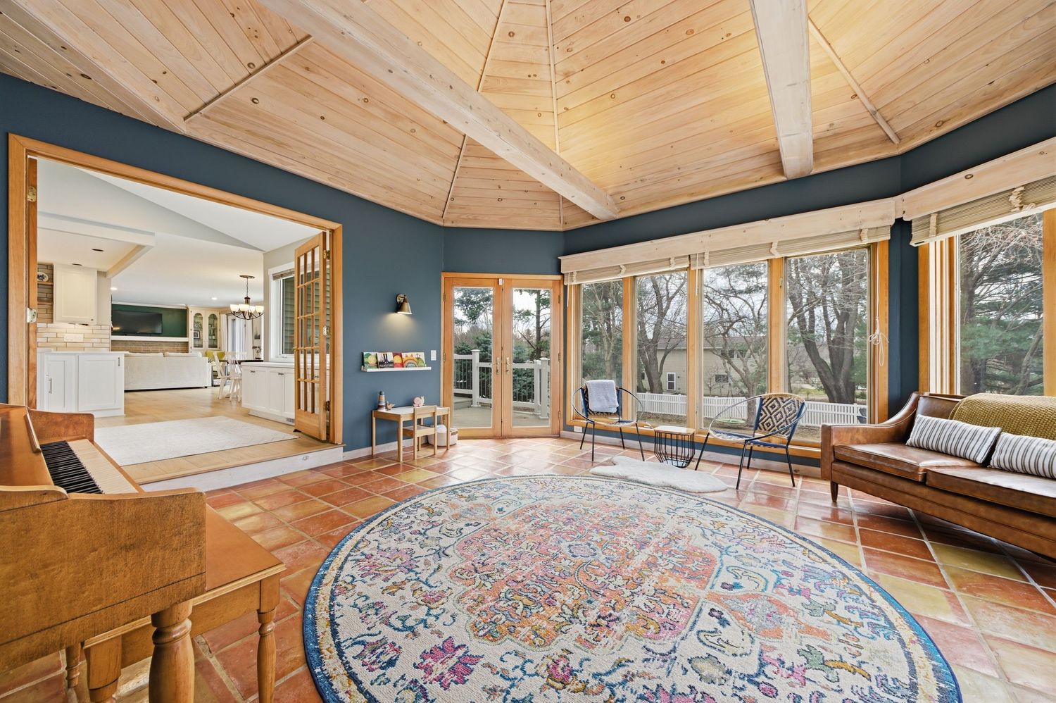 Sunlit four-season room with vaulted wood ceiling, terracotta tile floors, colorful round rug, and large windows overlooking trees.