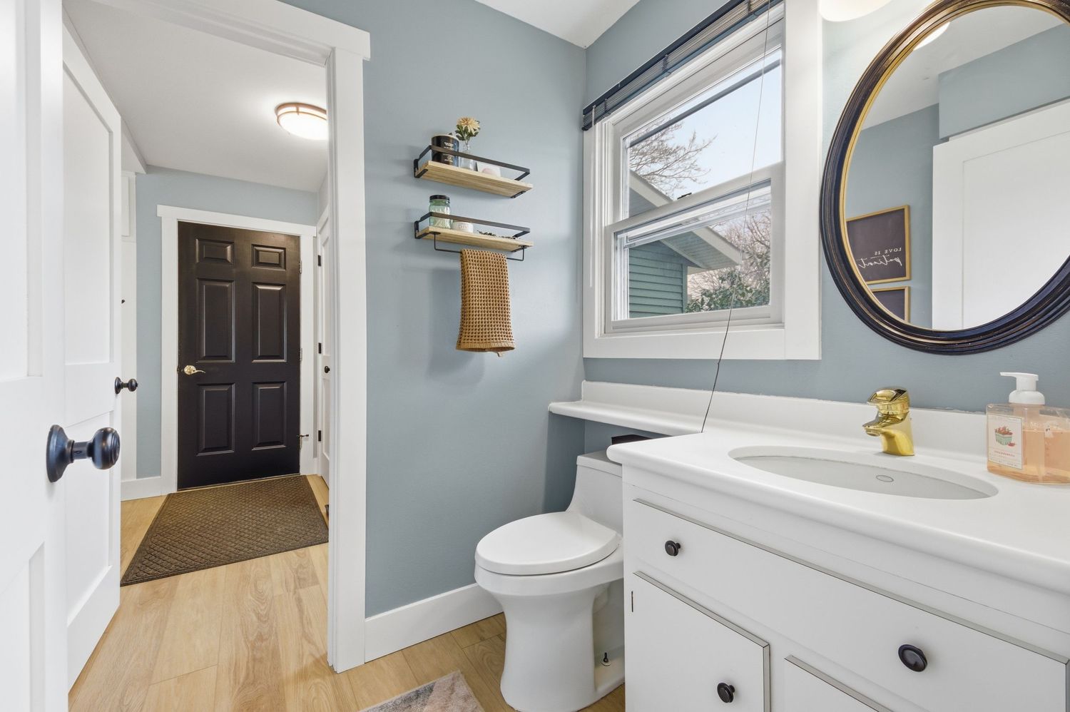 Modern bathroom with white vanity, round mirror, floating shelves, black door, and light blue walls with natural wood flooring.
