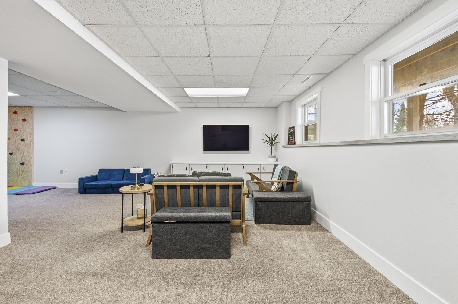 Finished basement rec room with carpet, drop ceiling, wall-mounted TV, blue sofa, and natural light from windows.