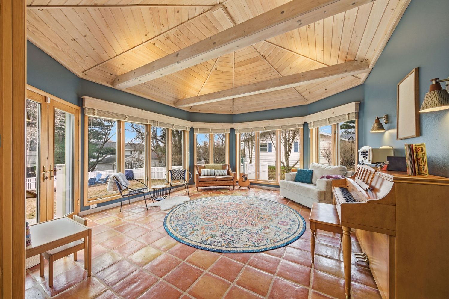 Sunlit sunroom with terracotta tile floors, vaulted wood ceiling, large windows, and cozy seating around a colorful round rug.